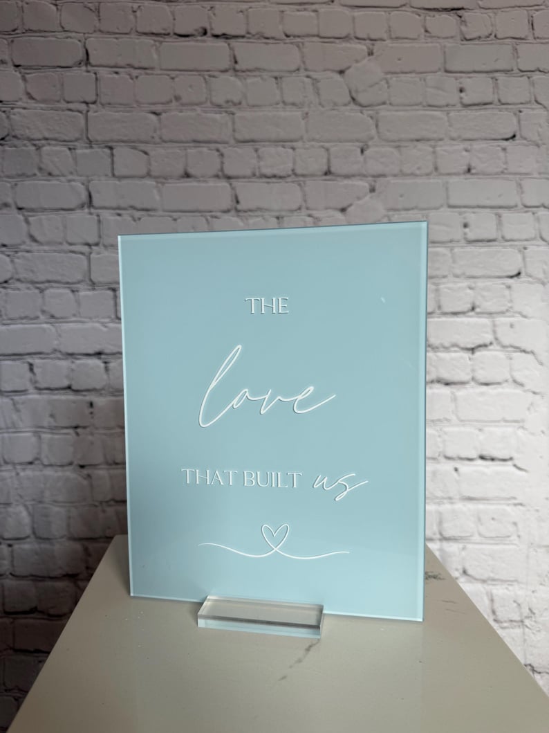 May include: A light blue acrylic sign with the words "THE love THAT BUILT us" in white script. A white heart is drawn below the text. The sign is displayed on a clear acrylic stand.