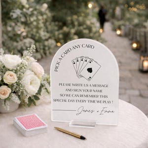 May include: An arched clear acrylic sign with black text reading PICK A CARD ANY CARD, PLEASE WRITE US A MESSAGE AND SIGN YOUR NAME SO WE CAN REMEMBER THIS SPECIAL DAY EVERY TIME WE PLAY!, and James + Emma, featuring a playing card graphic.