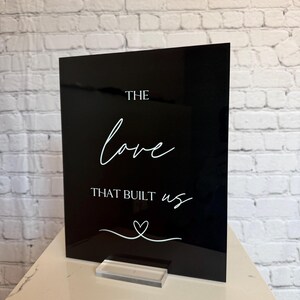 May include: A black acrylic sign with white script that reads "THE love THAT BUILT us" with a heart symbol. The sign is displayed on a clear acrylic stand, set against a white brick wall.