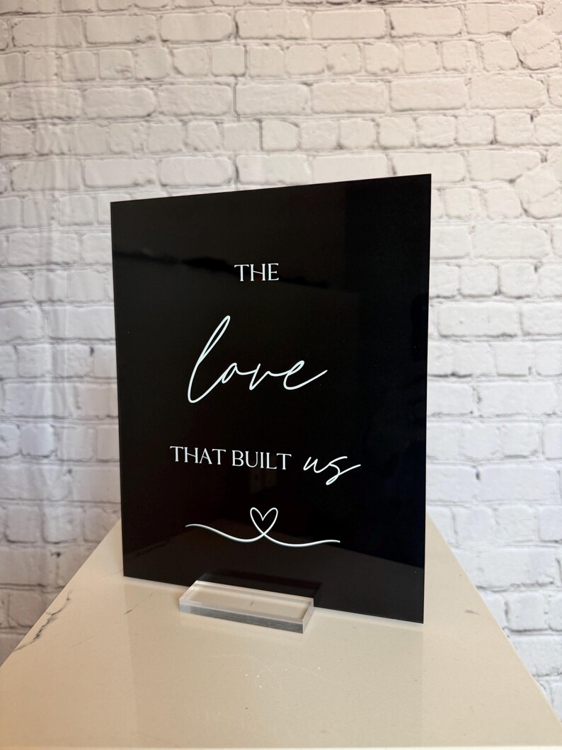 May include: A black acrylic sign with white script that reads "THE love THAT BUILT us" with a heart symbol. The sign is displayed on a clear acrylic stand, set against a white brick wall.