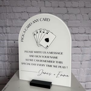 May include: An arched white acrylic sign with black text and a playing card graphic. The text reads: PICK A CARD ANY CARD, PLEASE WRITE US A MESSAGE AND SIGN YOUR NAME SO WE CAN REMEMBER THIS SPECIAL DAY EVERY TIME WE PLAY! James + Emma.