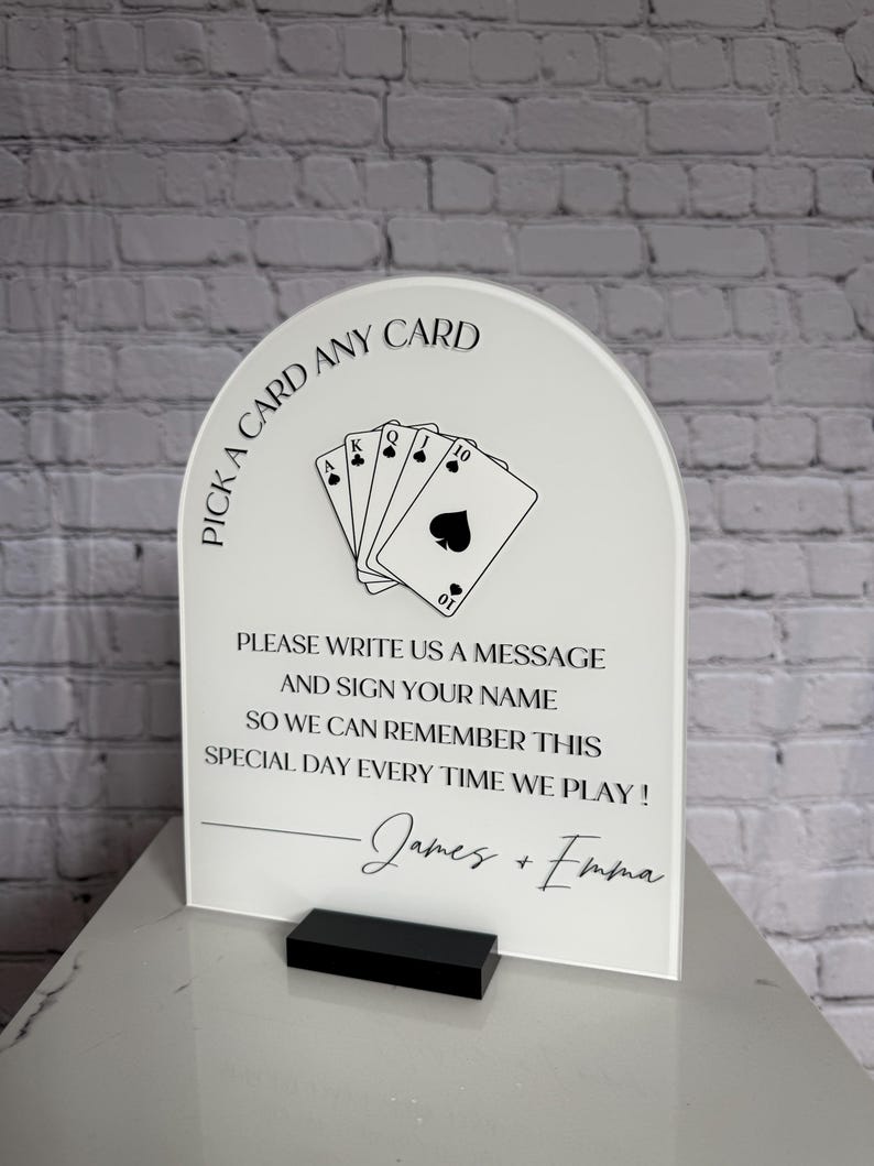 May include: An arched white acrylic sign with black text and a playing card graphic. The text reads: PICK A CARD ANY CARD, PLEASE WRITE US A MESSAGE AND SIGN YOUR NAME SO WE CAN REMEMBER THIS SPECIAL DAY EVERY TIME WE PLAY! James + Emma.