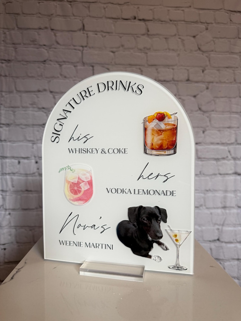 May include: A white acrylic sign with an arched top, displaying "SIGNATURE DRINKS." It details "his" Whiskey & Coke, "hers" Vodka Lemonade, and "Nova's" Weenie Martini, accompanied by drink illustrations and a black dog graphic.