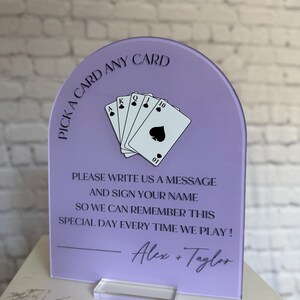 May include: An arched lavender acrylic sign featuring a black graphic of five playing cards and the text: PICK A CARD ANY CARD, PLEASE WRITE US A MESSAGE AND SIGN YOUR NAME SO WE CAN REMEMBER THIS SPECIAL DAY EVERY TIME WE PLAY! Alex + Taylor.