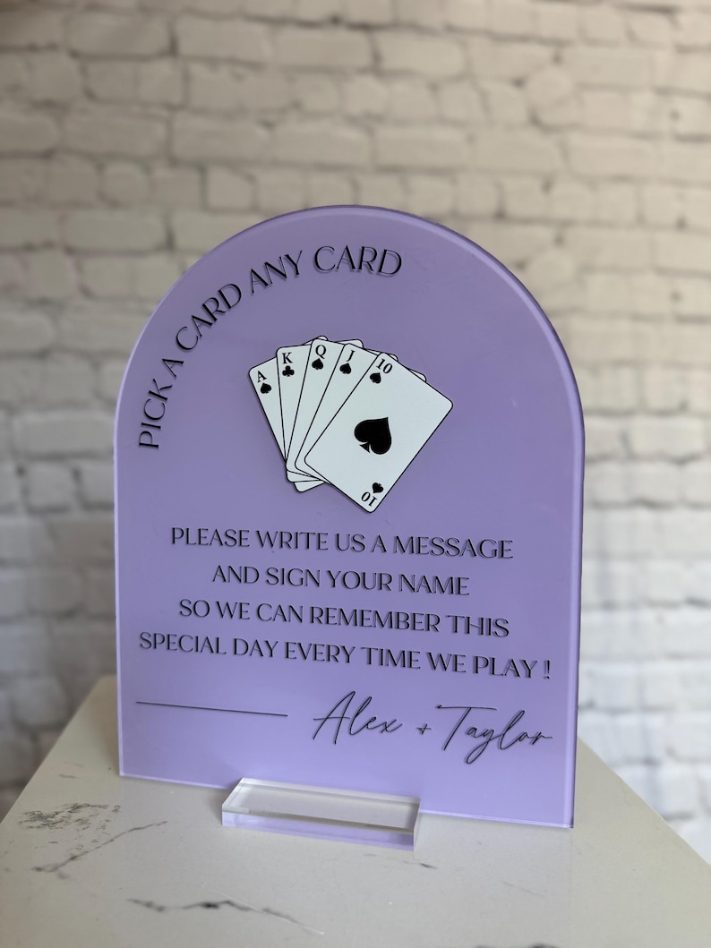May include: An arched lavender acrylic sign featuring a black graphic of five playing cards and the text: PICK A CARD ANY CARD, PLEASE WRITE US A MESSAGE AND SIGN YOUR NAME SO WE CAN REMEMBER THIS SPECIAL DAY EVERY TIME WE PLAY! Alex + Taylor.