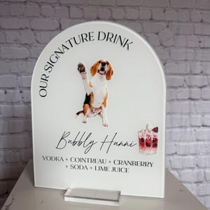 May include: A clear acrylic sign with a white background. The sign features a cartoon beagle dog waving, the text "OUR SIGNATURE DRINK", and the words "Bubbly Huani" in cursive. Below the text are the ingredients for the drink.
