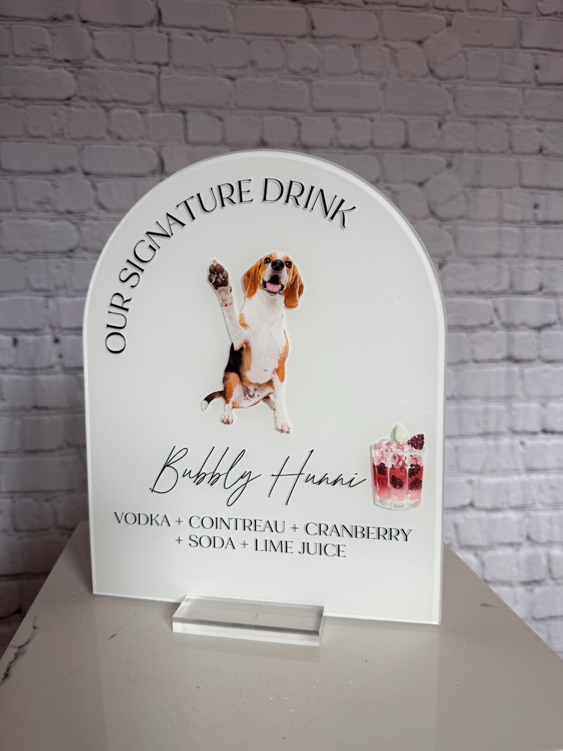 May include: A clear acrylic sign with a white background. The sign features a cartoon beagle dog waving, the text "OUR SIGNATURE DRINK", and the words "Bubbly Huani" in cursive. Below the text are the ingredients for the drink.
