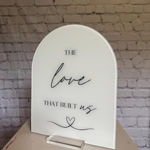 May include: A white acrylic sign with an arched top. The sign features the words "THE love THAT BUILT us" in black script, along with a heart symbol. It stands on a clear acrylic base.