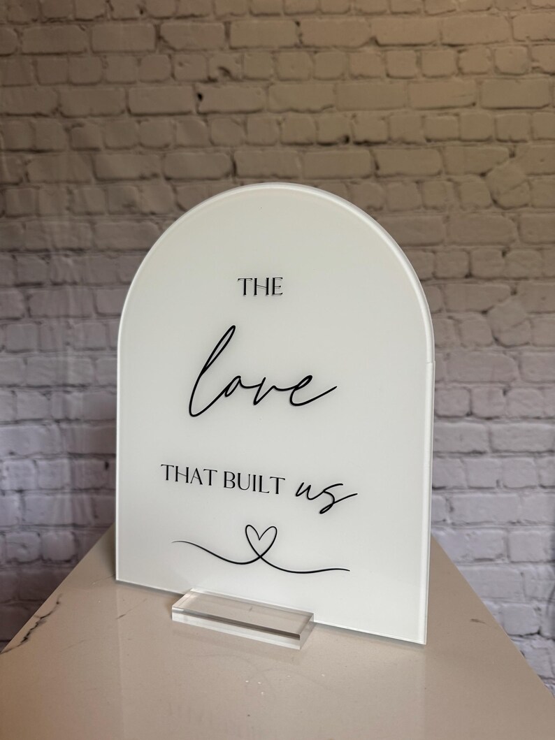 May include: A white acrylic sign with an arched top. The sign features the words "THE love THAT BUILT us" in black script, along with a heart symbol. It stands on a clear acrylic base.