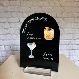 Może przedstawiać: Czarny akrylowy znak z napisem "Signature Drinks" i ilustracjami koktajli. Zawiera "his" z Whiskey Sour i "hers" z French 75. Przezroczysta akrylowa podstawa wspiera znak.