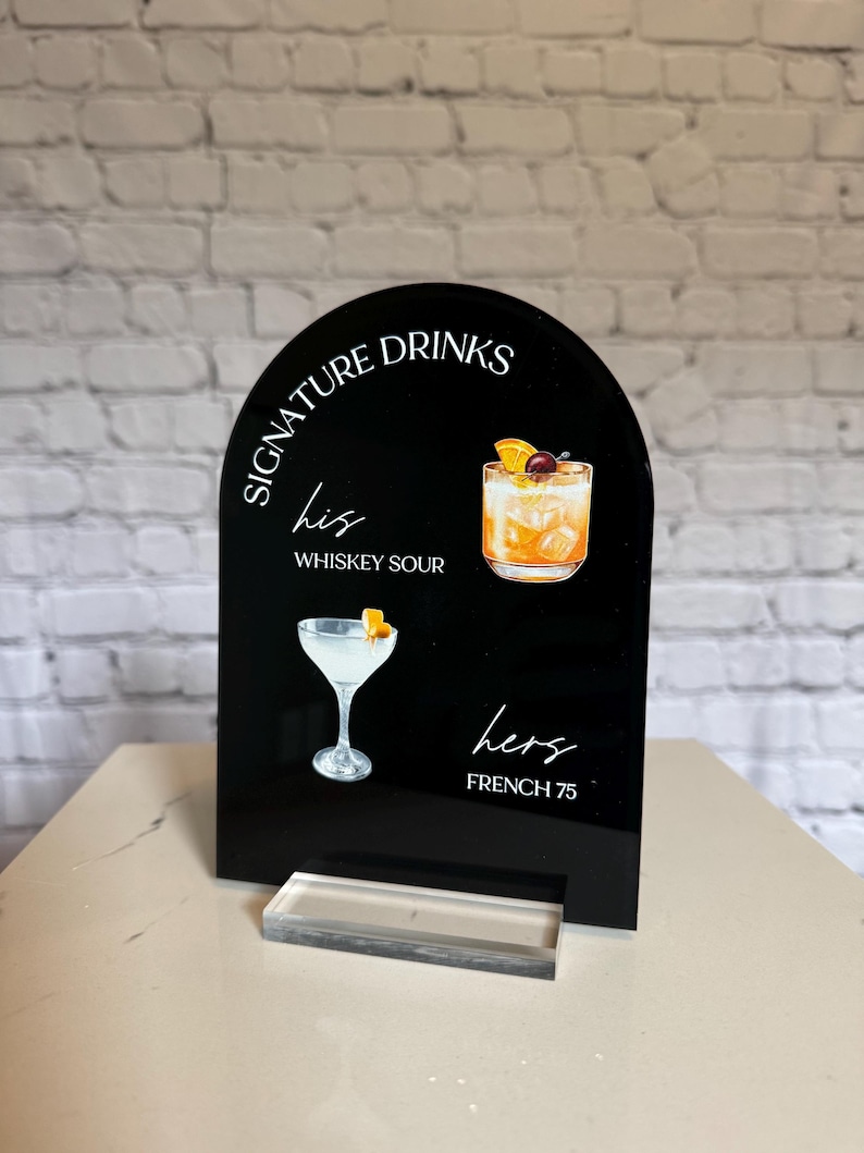 Może przedstawiać: Czarny akrylowy znak z napisem "Signature Drinks" i ilustracjami koktajli. Zawiera "his" z Whiskey Sour i "hers" z French 75. Przezroczysta akrylowa podstawa wspiera znak.