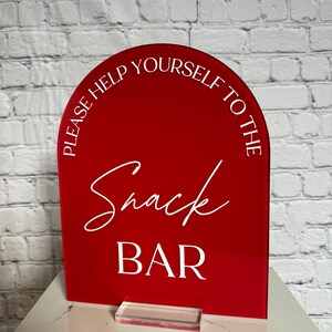 May include: A red arched acrylic sign with white text that reads "PLEASE HELP YOURSELF TO THE" in a sans-serif font, "Snack" in a cursive script, and "BAR" in a bold sans-serif font. The sign is supported by a clear rectangular acrylic base.