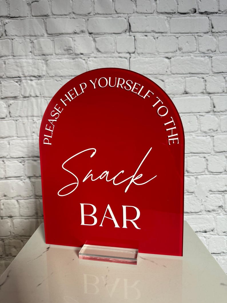 May include: A red arched acrylic sign with white text that reads "PLEASE HELP YOURSELF TO THE" in a sans-serif font, "Snack" in a cursive script, and "BAR" in a bold sans-serif font. The sign is supported by a clear rectangular acrylic base.