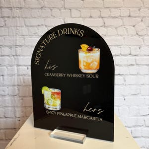Może przedstawiać: Czarny akrylowy znak z napisem "Signature Drinks" i ilustracjami koktajli. Znak zawiera tekst "his" i "hers" z nazwami drink&oacute;w: Cranberry Whiskey Sour i Spicy Pineapple Margarita.