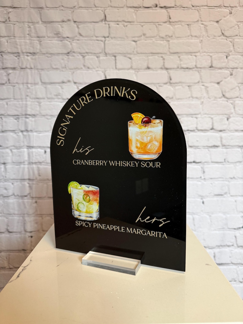 Może przedstawiać: Czarny akrylowy znak z napisem "Signature Drinks" i ilustracjami koktajli. Znak zawiera tekst "his" i "hers" z nazwami drink&oacute;w: Cranberry Whiskey Sour i Spicy Pineapple Margarita.