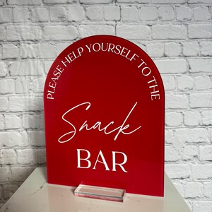 May include: A red arched acrylic sign with white text that reads PLEASE HELP YOURSELF TO THE Snack BAR. The sign is mounted on a clear rectangular acrylic base, featuring a smooth, glossy finish and a clean, modern design for event signage.