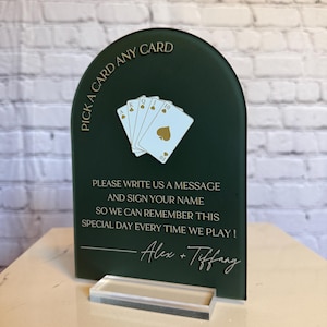 May include: An arched dark green sign with gold text reading PICK A CARD ANY CARD and PLEASE WRITE US A MESSAGE AND SIGN YOUR NAME SO WE CAN REMEMBER THIS SPECIAL DAY EVERY TIME WE PLAY! with Alex + Tiffany, featuring a fan of four playing cards.