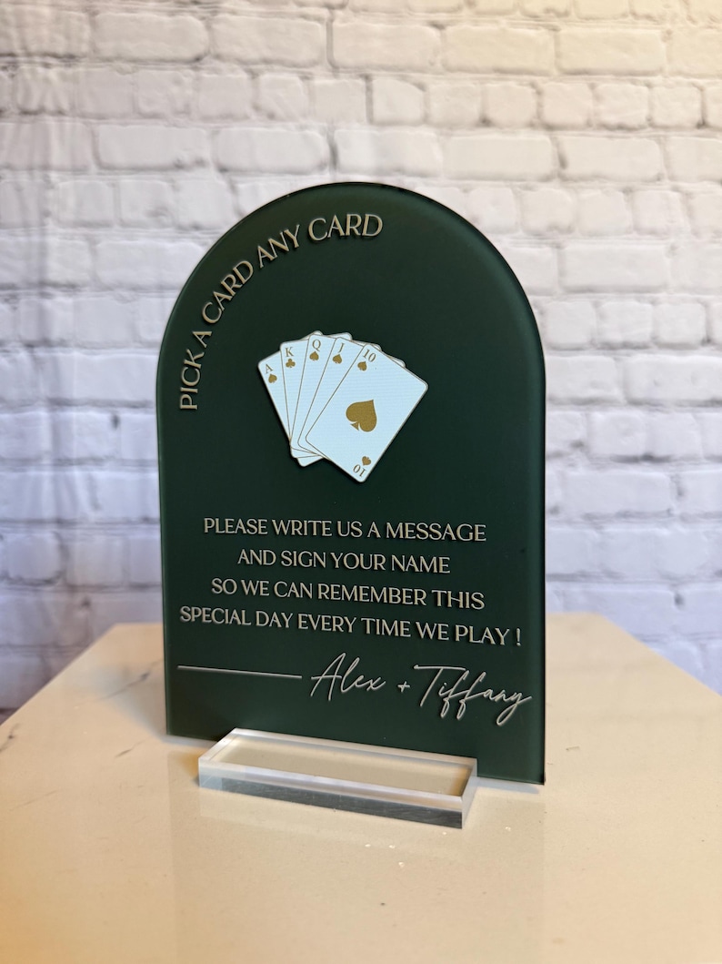 May include: An arched dark green sign with gold text reading PICK A CARD ANY CARD and PLEASE WRITE US A MESSAGE AND SIGN YOUR NAME SO WE CAN REMEMBER THIS SPECIAL DAY EVERY TIME WE PLAY! with Alex + Tiffany, featuring a fan of four playing cards.