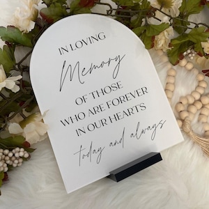 May include: A white acrylic memorial sign with a rounded top, inscribed with the words "IN LOVING Memory OF THOSE WHO ARE FOREVER IN OUR HEARTS today and always" in black script. The sign is on a black stand, with floral and beaded decorations.