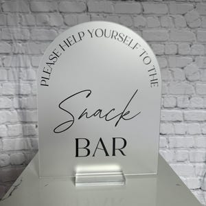 May include: An arched, frosted acrylic sign with a clear base. The text in black reads "PLEASE HELP YOURSELF TO THE" in a sans-serif font, followed by the word "Snack" in a cursive script, and the word "BAR" in a bold, capitalized sans-serif font.