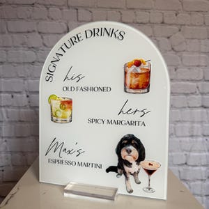 May include: White acrylic drinks sign featuring cocktail illustrations and the text "Signature Drinks". The sign includes the text "his Old Fashioned", "hers Spicy Margarita", and "Max's Espresso Martini". A dog illustration is also featured.