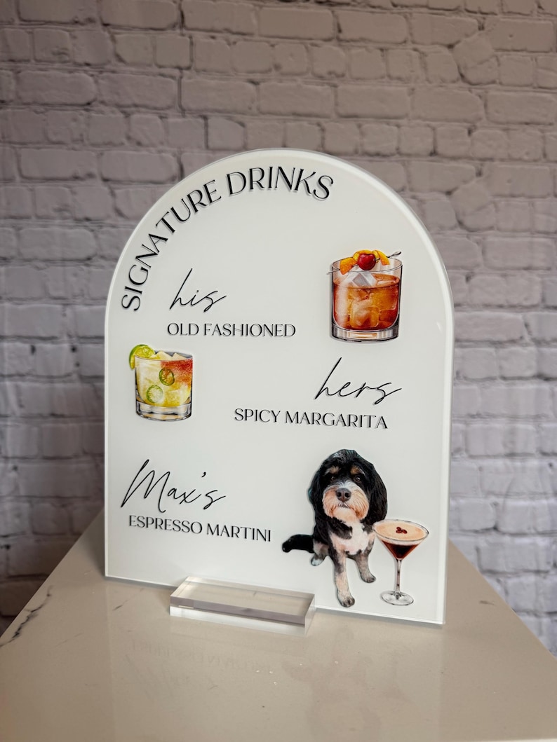 May include: White acrylic drinks sign featuring cocktail illustrations and the text "Signature Drinks". The sign includes the text "his Old Fashioned", "hers Spicy Margarita", and "Max's Espresso Martini". A dog illustration is also featured.