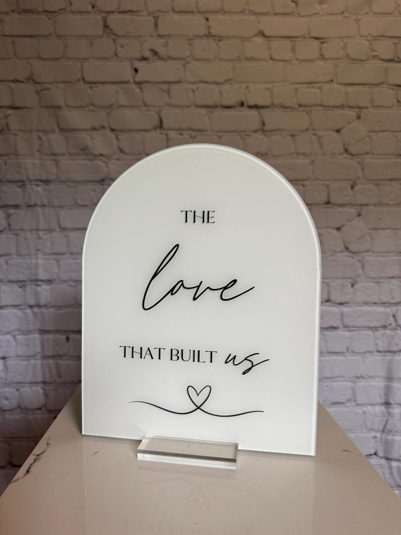 May include: White arched sign with the text "THE love THAT BUILT us" in black script. A heart graphic is below the text. The sign is displayed on a clear acrylic stand.
