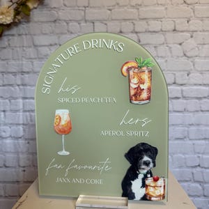 May include: A sage green acrylic drinks sign with the words "Signature Drinks" and drink illustrations. The sign features "His" Spiced Peach Tea, "Hers" Aperol Spritz, and "fan favourite" Jaxx and Coke. A black and white dog is also featured.