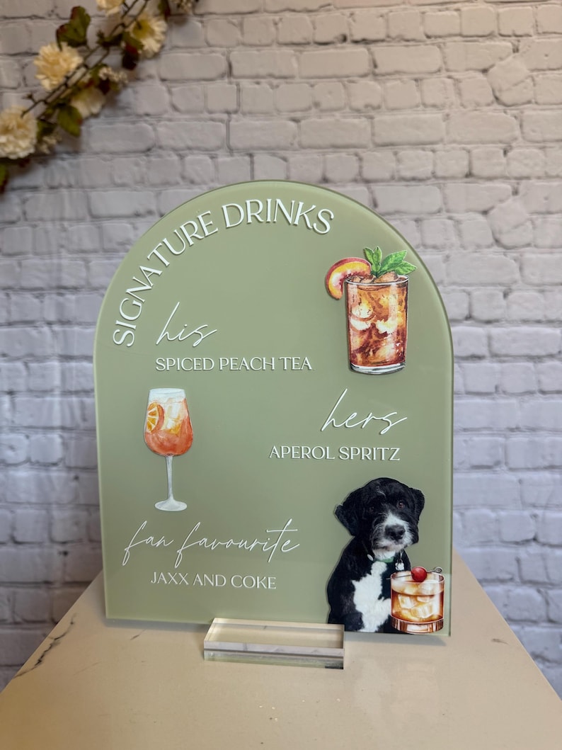May include: A sage green acrylic drinks sign with the words "Signature Drinks" and drink illustrations. The sign features "His" Spiced Peach Tea, "Hers" Aperol Spritz, and "fan favourite" Jaxx and Coke. A black and white dog is also featured.