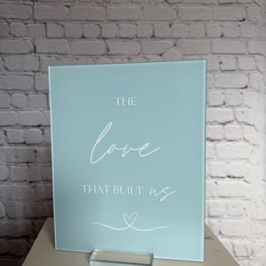 May include: A square, light blue sign with the words "THE love THAT BUILT us" in white script. A white heart is drawn below the text. The sign is displayed on a clear acrylic stand.