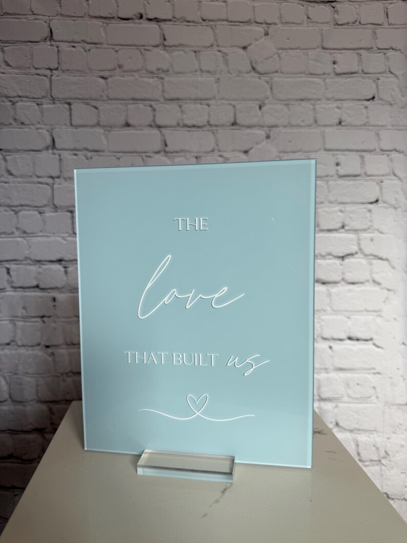 May include: A square, light blue sign with the words "THE love THAT BUILT us" in white script. A white heart is drawn below the text. The sign is displayed on a clear acrylic stand.