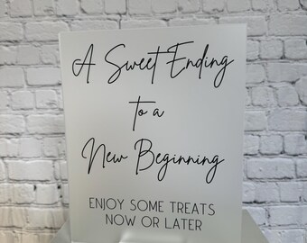 May include: A clear acrylic sign with black text that reads "A Sweet Ending to a New Beginning. Enjoy some treats now or later."