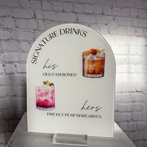 Może przedstawiać: Biały akrylowy znak z łukowatym wierzchołkiem, z napisem "Signature Drinks". Zawiera ilustracje koktajli: Old Fashioned i Prickly Pear Margarita. Znak stoi na przezroczystej akrylowej podstawie.