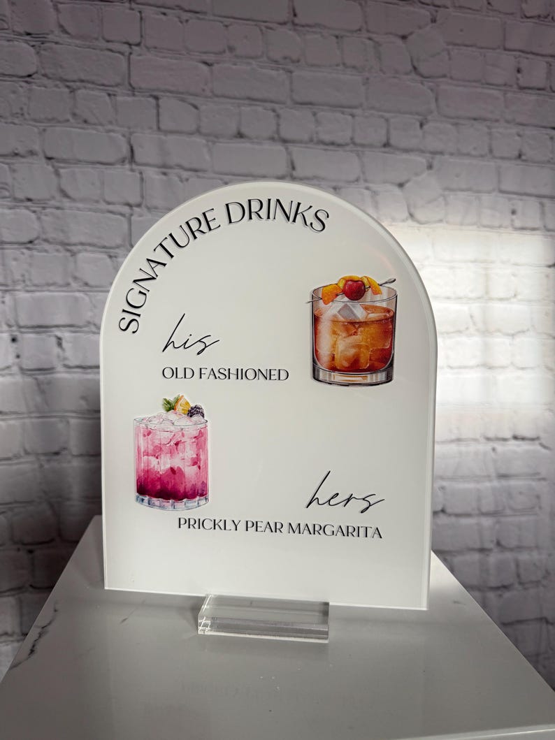 Może przedstawiać: Biały akrylowy znak z łukowatym wierzchołkiem, z napisem "Signature Drinks". Zawiera ilustracje koktajli: Old Fashioned i Prickly Pear Margarita. Znak stoi na przezroczystej akrylowej podstawie.