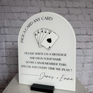 May include: An arched white acrylic sign with black text and a graphic of five playing cards. The text reads: PICK A CARD ANY CARD, PLEASE WRITE US A MESSAGE AND SIGN YOUR NAME SO WE CAN REMEMBER THIS SPECIAL DAY EVERY TIME WE PLAY! James + Emma.