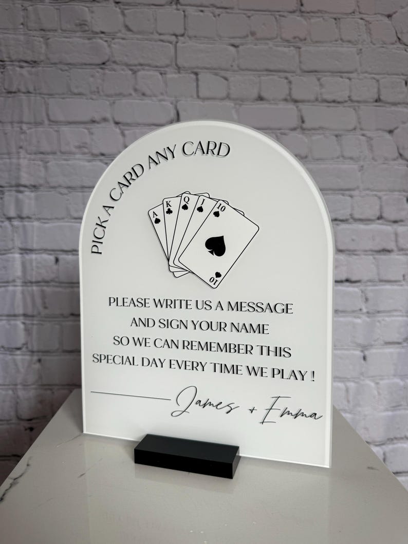 May include: An arched white acrylic sign with black text and a graphic of five playing cards. The text reads: PICK A CARD ANY CARD, PLEASE WRITE US A MESSAGE AND SIGN YOUR NAME SO WE CAN REMEMBER THIS SPECIAL DAY EVERY TIME WE PLAY! James + Emma.