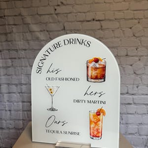 May include: White acrylic sign with an arched top, featuring the words "Signature Drinks." It displays illustrations of cocktails: an Old Fashioned, Dirty Martini, and Tequila Sunrise. The sign includes the words "his," "hers," and "ours."