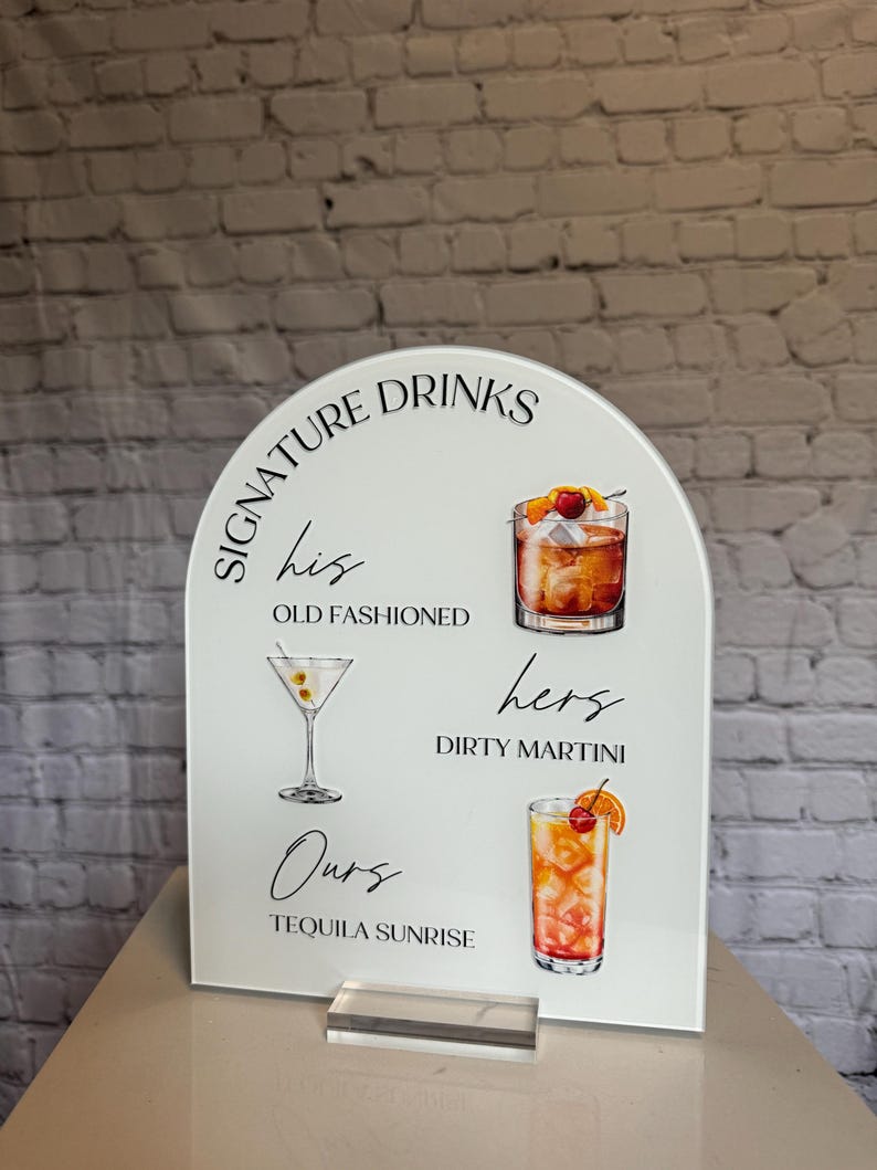 May include: White acrylic sign with an arched top, featuring the words "Signature Drinks." It displays illustrations of cocktails: an Old Fashioned, Dirty Martini, and Tequila Sunrise. The sign includes the words "his," "hers," and "ours."