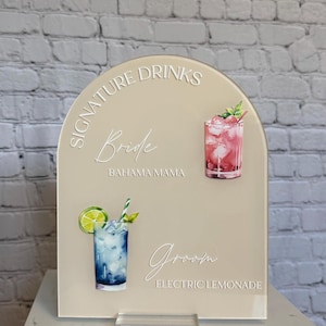Może przedstawiać: Beżowy akrylowy znak z łukowatym wierzchem, z napisami "Signature Drinks" oraz imionami "Bride" i "Groom". Zawiera akwarelowe ilustracje dw&oacute;ch koktajli: r&oacute;żowego napoju i niebieskiego napoju z limonką. Znak jest zamontowany na przezroczystej akrylowej podstawie.