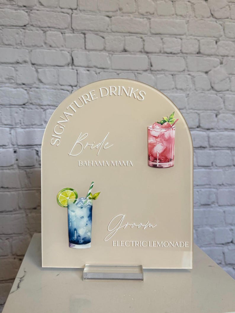 Może przedstawiać: Beżowy akrylowy znak z łukowatym wierzchem, z napisami "Signature Drinks" oraz imionami "Bride" i "Groom". Zawiera akwarelowe ilustracje dw&oacute;ch koktajli: r&oacute;żowego napoju i niebieskiego napoju z limonką. Znak jest zamontowany na przezroczystej akrylowej podstawie.