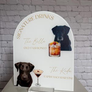 May include: A white acrylic sign with gold lettering displaying "Signature Drinks." The sign features illustrations of cocktails and black Labrador dogs. The cocktails are "The Bella Old Fashioned" and "The Koda Espresso Martini."