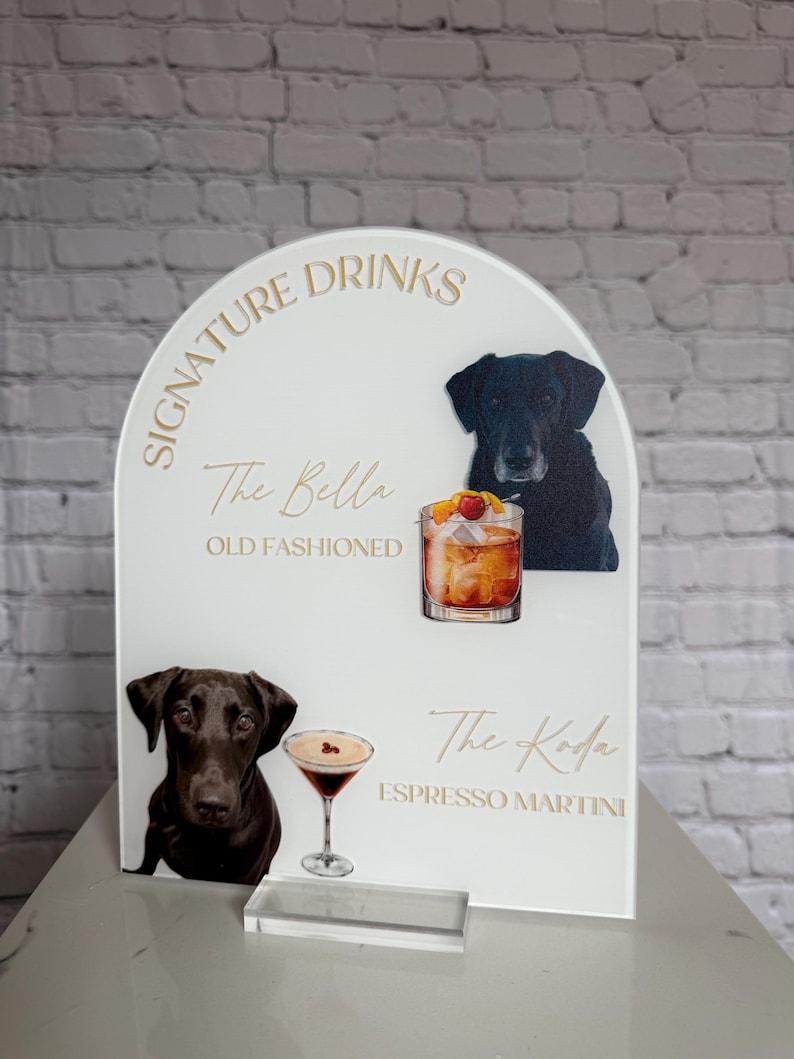 May include: A white acrylic sign with gold lettering displaying "Signature Drinks." The sign features illustrations of cocktails and black Labrador dogs. The cocktails are "The Bella Old Fashioned" and "The Koda Espresso Martini."