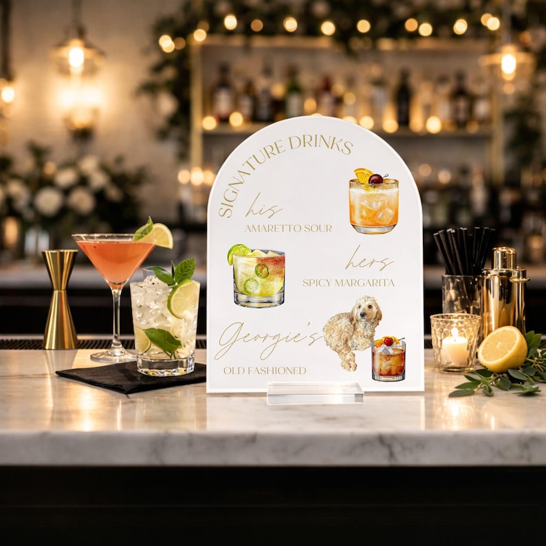 May include: A white, arched sign with gold lettering displaying "Signature Drinks". The sign features cocktail illustrations and a dog, with text including "Amaretto Sour", "Spicy Margarita", and "Georgie's Old Fashioned".