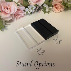 May include: Clear and black acrylic stand options are displayed, with the text "Stand Options" at the bottom. The clear acrylic stands are rectangular, and the black acrylic stands are also rectangular. Floral arrangements in pink and white are in the background.