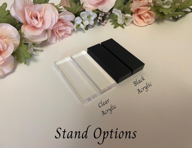 May include: Clear and black acrylic stand options are displayed, with the text "Stand Options" at the bottom. The clear acrylic stands are rectangular, and the black acrylic stands are also rectangular. Floral arrangements in pink and white are in the background.