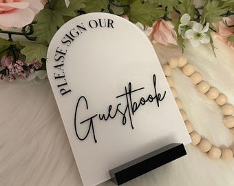 May include: White acrylic sign with black text that reads "Please sign our Guestbook". The sign is shaped like an arch and is standing on a black stand.