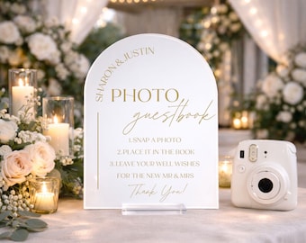 May include: An arched white acrylic sign with gold text reading SHARON & JUSTIN PHOTO guestbook, 1. SNAP A PHOTO, 2. PLACE IT IN THE BOOK, 3. LEAVE YOUR WELL WISHES FOR THE NEW MR & MRS, Thank You!, positioned next to a white instant film camera.