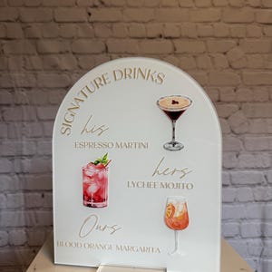 May include: A white acrylic sign with an arched top, featuring the words "Signature Drinks" and three cocktail illustrations: Espresso Martini, Lychee Mojito, and Blood Orange Margarita. The sign is displayed on a clear acrylic stand.