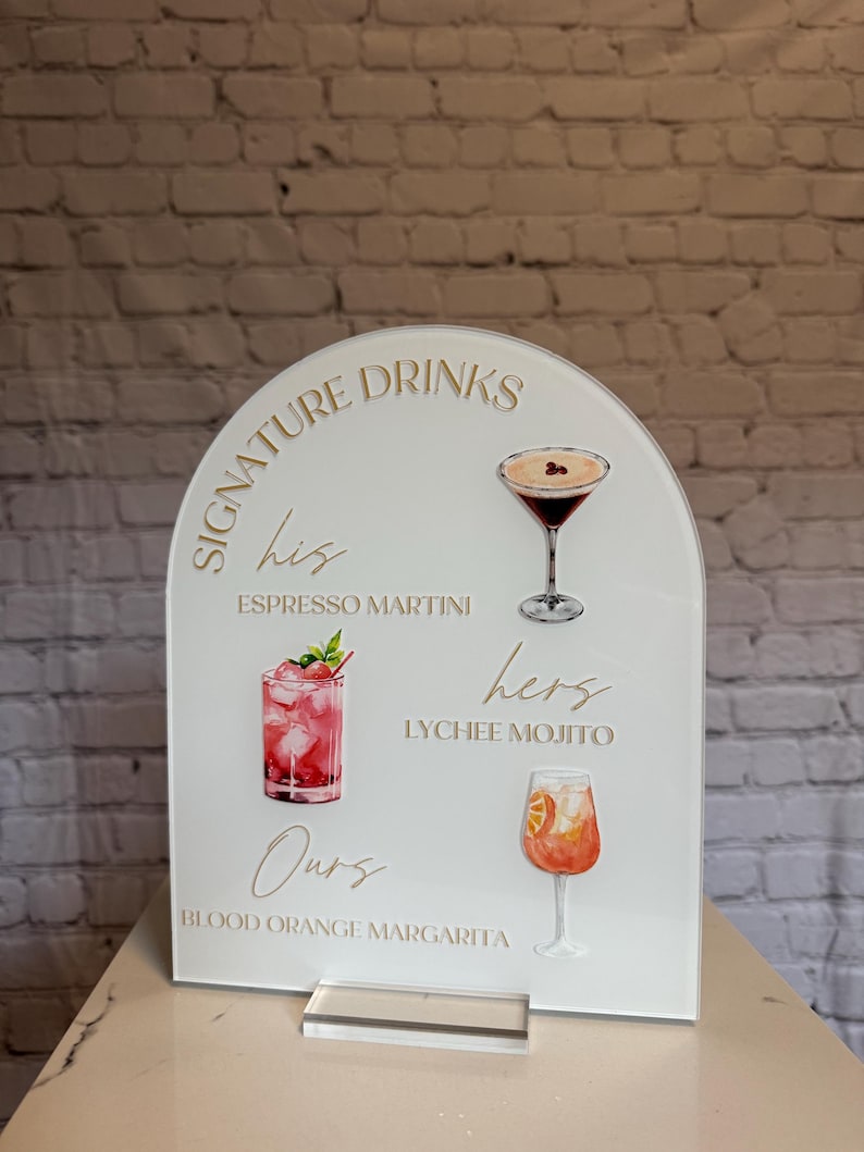 May include: A white acrylic sign with an arched top, featuring the words "Signature Drinks" and three cocktail illustrations: Espresso Martini, Lychee Mojito, and Blood Orange Margarita. The sign is displayed on a clear acrylic stand.