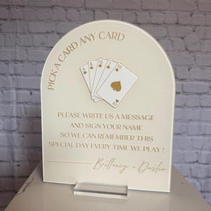 May include: An arched cream-colored acrylic sign featuring gold text and a graphic of five playing cards. The text reads: PICK A CARD ANY CARD, PLEASE WRITE US A MESSAGE AND SIGN YOUR NAME SO WE CAN REMEMBER THIS SPECIAL DAY EVERY TIME WE PLAY! Brittany + Dustin.